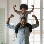 Small son sit on strong dad shoulders showing biceps. African family enjoy activity games at home, healthy fit lifestyle, two superheroes, vitamins for adults and children ad, happy Father Day concept